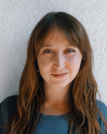Eine Frau mit langen, braunen Haaren und einem Nasenring schaut direkt in die Kamera. Sie trägt ein graues T-Shirt und hat einen neutralen Gesichtsausdruck. Der Hintergrund ist hell und einfarbig.