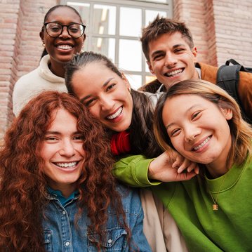 Five young people are smiling happily at the camera as they pose together. They are wearing casual clothes and standing in front of a large window.