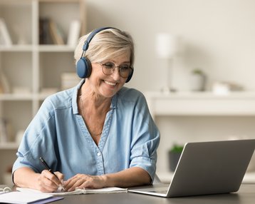 Eine ältere Frau mit Kopfhörern lächelt, während sie an einem Laptop arbeitet und Notizen macht.