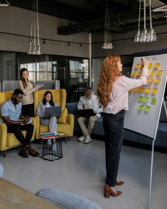 Eine Gruppe von Menschen in einem modernen Büro, die an einem Meeting teilnehmen. Eine Frau mit langen, roten Haaren schreibt auf ein Flipchart, während andere Teilnehmer Notizen machen oder auf Laptops arbeiten.