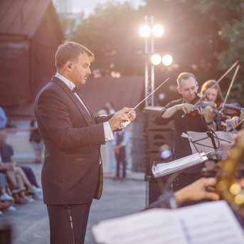 A conductor leads an orchestra outdoors while musicians play string and wind instruments.