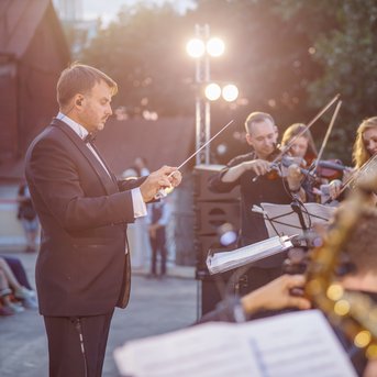Ein Dirigent leitet ein Orchester im Freien, während Musiker mit Streichinstrumenten und Blasinstrumenten spielen.