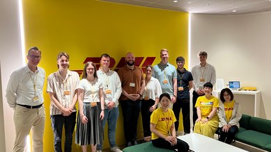 Eine Gruppe von zwölf Personen steht in einem modernen Raum mit gelber Wand und DHL-Logo, alle tragen Namensschilder.