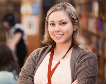 A smiling woman stands in a library, surrounded by bookshelves. In the background, other people can be seen working on computers or chatting with each other.