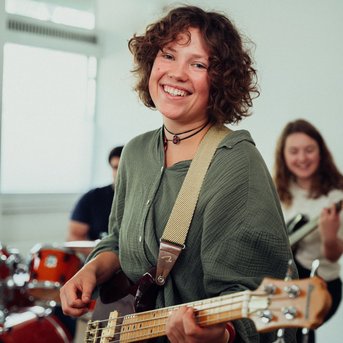 Eine Frau mit lockigem Haar spielt Bass in einer Bandprobe, lächelnd und in einem lässigen Outfit. Im Hintergrund sind Schlagzeug und andere Musiker sichtbar.