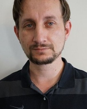 Ein Mann mit kurzen, braunen Haaren und einem leichten Bart trägt ein schwarzes Polohemd mit grauen Streifen. Der Hintergrund ist neutral.