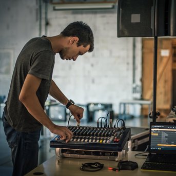 Ein junger Mann bedient ein Mischpult und einen Laptop in einem Raum mit minimalistischem Design.