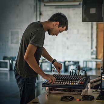 A young man is operating a mixing console and a laptop in a room with minimalist design.