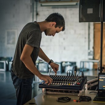 Ein junger Mann bedient ein Mischpult und einen Laptop in einem Raum mit minimalistischem Design.