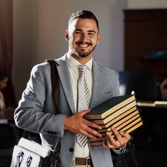 Ein junger Mann in einem Anzug lächelt, während er mehrere Bücher in den Händen hält. Im Hintergrund sind andere Personen in einem Büro zu sehen.