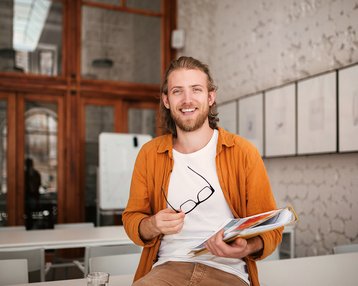 Ein junger Mann mit langen Haaren und Bart sitzt auf einem Tisch in einem modernen Raum. Er hält eine Mappe mit Dokumenten und eine Brille in der Hand. Er trägt ein oranges Hemd und ein weißes T-Shirt.