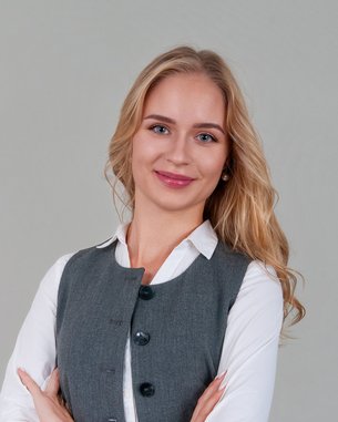 A woman with long, blonde hair is wearing a gray vest over a white shirt and is standing with her arms crossed in front of a neutral background.