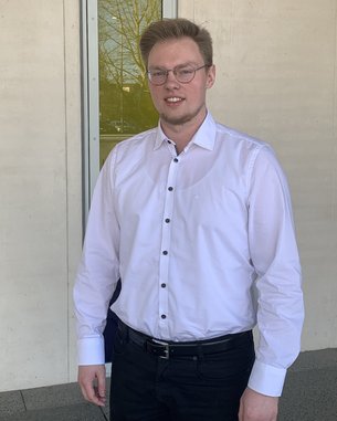 Ein junger Mann in einem weißen Hemd steht vor einer modernen Wand mit Fenstern. Er trägt eine Brille und hat kurzes, blondes Haar.