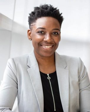 Eine lächelnde Frau mit kurzem, lockigem Haar trägt einen grauen Blazer über einem schwarzen Oberteil. Sie sitzt an einem Tisch und posiert in einem modernen, hellen Raum mit großen Fenstern.