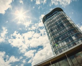 Ein modernes Hochhaus mit einer geschwungenen Fassade, das unter einem klaren Himmel mit Wolken und Sonnenschein steht.