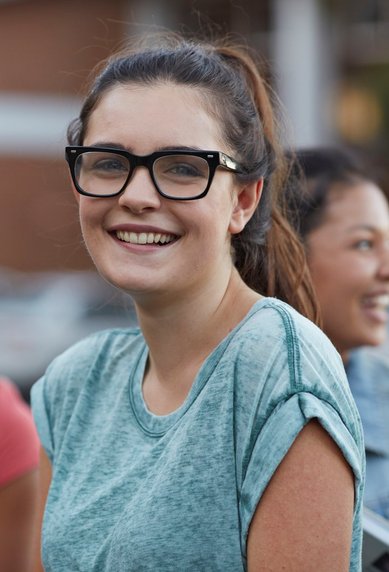 Eine Gruppe junger Menschen steht zusammen, während eine Frau mit Brille in der Mitte lächelt. Sie trägt ein graues T-Shirt und hat lockige Haare. Die anderen Personen scheinen ebenfalls freundlich zu sein.
