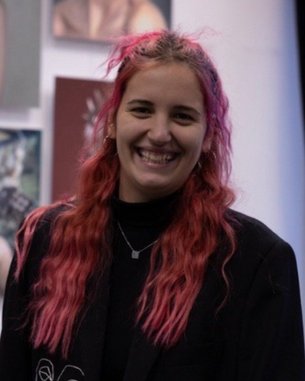 Eine Person mit langen, pinken Haaren lächelt in einem Raum mit Kunstwerken im Hintergrund. Sie trägt einen schwarzen Blazer und eine Kette.