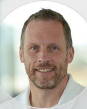 Ein Mann mit kurzem, grauem Haar und Bart lächelt in die Kamera. Er trägt ein weißes Hemd und hat einen neutralen Hintergrund.