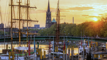 Hafenansicht bei Sonnenuntergang mit historischen Schiffen, einer Kirche im Hintergrund und Spaziergängern am Ufer.