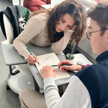A person is drawing in a notebook while another person points to the first person's hand with a pen.