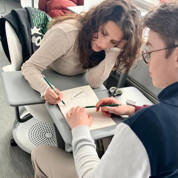 Eine Person zeichnet in ein Notizbuch, während eine andere Person mit einem Stift auf die Hand der ersten Person zeigt.