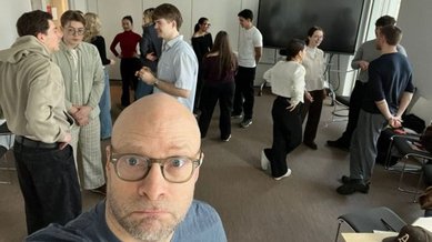 Eine Gruppe von Menschen steht in einem Raum und unterhält sich. Im Vordergrund ist ein Mann mit Brille zu sehen, der in die Kamera schaut.