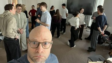 A group of people is standing in a room and having a conversation. In the foreground, there is a man with glasses looking at the camera.