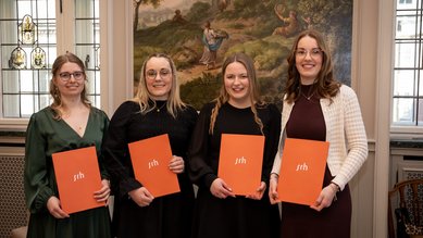 Vier Frauen halten orangefarbene Urkunden der SRH in einer festlichen Umgebung. Sie stehen nebeneinander und lächeln in die Kamera.
