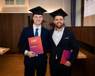 Zwei Männer in Graduationskleidung halten Urkunden in den Händen und lächeln. Der Hintergrund zeigt eine festliche Umgebung.