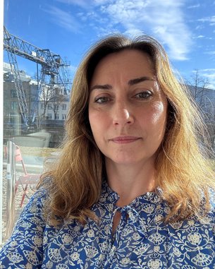 Frau mit langen, braunen Haaren und blauer Bluse, steht vor einem Fenster mit einem Blick auf eine Baustelle und einen klaren Himmel.