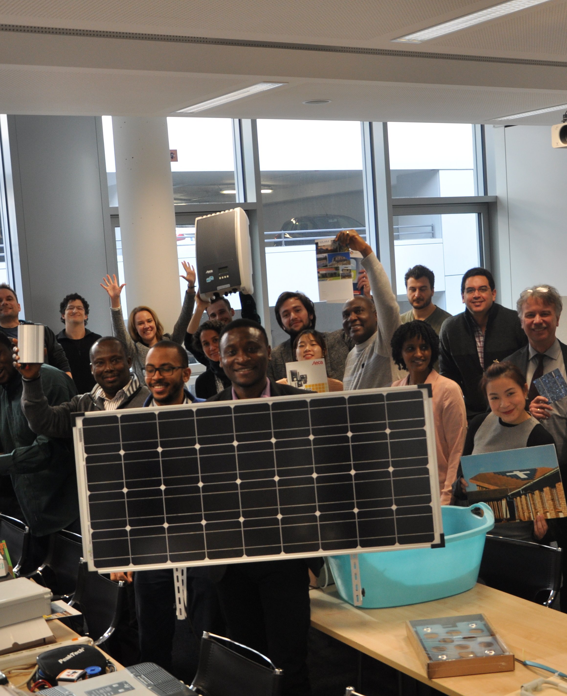 Eine Gruppe von Menschen steht in einem Raum und hält verschiedene Gegenstände, darunter ein Solarpanel. Sie lächeln und posieren für das Foto, während einige mit den Händen Winken oder Daumen hoch zeigen.