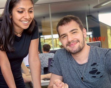 Two people in a modern office, a woman is standing smiling next to a man who is working at the computer.