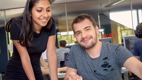 Two people in a modern office, a woman is standing smiling next to a man who is working at the computer.