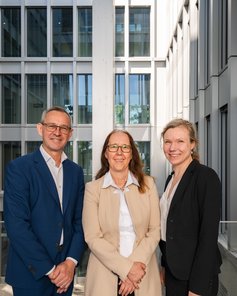 Drei Personen stehen in einem modernen Gebäude mit großen Fenstern. Sie lächeln und tragen formelle Kleidung.