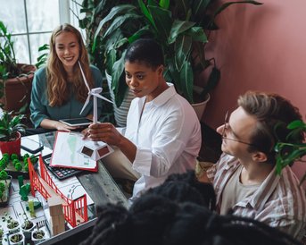A group of three people is discussing at a table surrounded by plants and a model of a wind turbine.