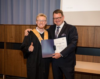 Ein graduierter Student in Robe hält ein Diplom und zeigt Daumen hoch, während er mit einem Mann in Anzug posiert.