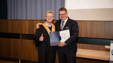 Ein graduierter Student in Robe hält ein Diplom und zeigt Daumen hoch, während er mit einem Mann in Anzug posiert.