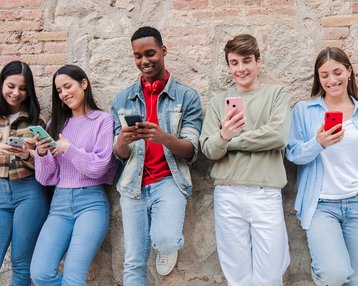 Five teenagers are standing by a wall, using their smartphones while interacting with each other.