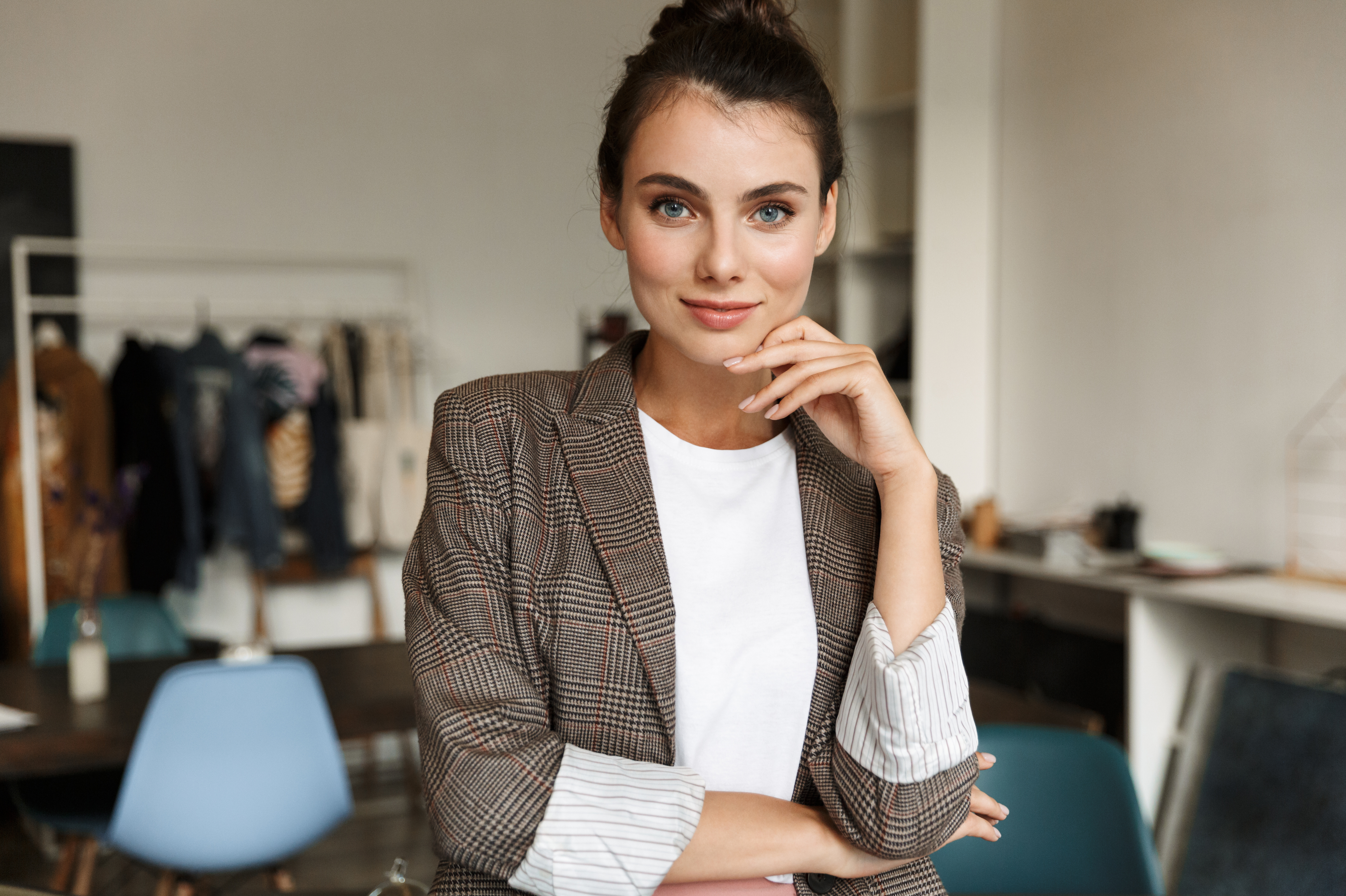Eine junge Frau mit braunem kariertem Blazer und weißem T-Shirt steht in einem modernen Raum. Sie hat einen selbstbewussten Ausdruck und hält eine Hand an ihrem Kinn. Im Hintergrund sind Möbel und Kleidung sichtbar.