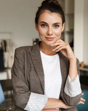 Eine junge Frau mit braunem kariertem Blazer und weißem T-Shirt steht in einem modernen Raum. Sie hat einen selbstbewussten Ausdruck und hält eine Hand an ihrem Kinn. Im Hintergrund sind Möbel und Kleidung sichtbar.