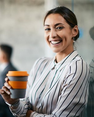 Eine lächelnde Frau mit gestreiftem Hemd hält einen Kaffeebecher in der Hand. Im Hintergrund sind zwei Personen in einem Besprechungsraum zu sehen. Die Atmosphäre wirkt professionell und modern.