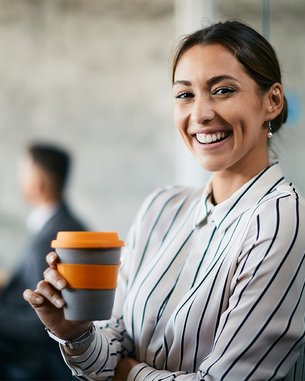 Eine lächelnde Frau mit gestreiftem Hemd hält einen Kaffeebecher in der Hand. Im Hintergrund sind zwei Personen in einem Besprechungsraum zu sehen. Die Atmosphäre wirkt professionell und modern.