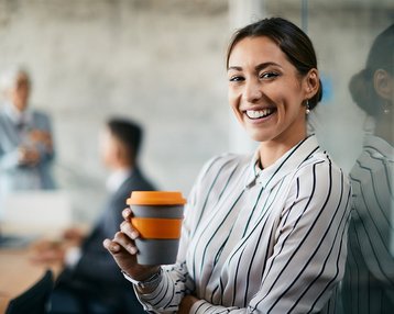 Eine lächelnde Frau mit gestreiftem Hemd hält einen Kaffeebecher in der Hand. Im Hintergrund sind zwei Personen in einem Besprechungsraum zu sehen. Die Atmosphäre wirkt professionell und modern.