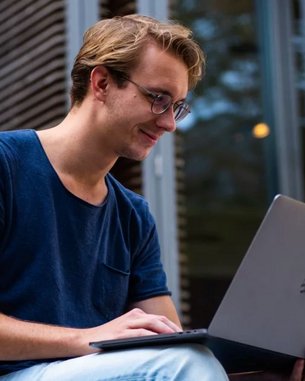 A young man is sitting outside, working on a laptop. He’s wearing a blue T-shirt and glasses as he focuses intently on the screen. In the background, trees and windows can be seen.