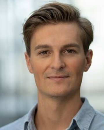 Ein junger Mann mit kurzen, gestylten Haaren und einem hellblauen Hemd schaut direkt in die Kamera. Der Hintergrund ist unscharf.