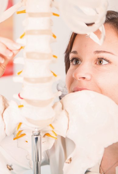 Eine Person untersucht ein anatomisches Skelettmodell, während sie eine Wirbelsäule anpasst. Die Umgebung ist hell und zeigt medizinische Illustrationen im Hintergrund. Die Person trägt eine helle Bluse und hat braune Haare.