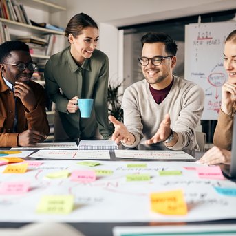 Vier Personen sitzen um einen Tisch, diskutieren und arbeiten an einem Projekt mit Notizen und Diagrammen. Eine Person hält eine Tasse.