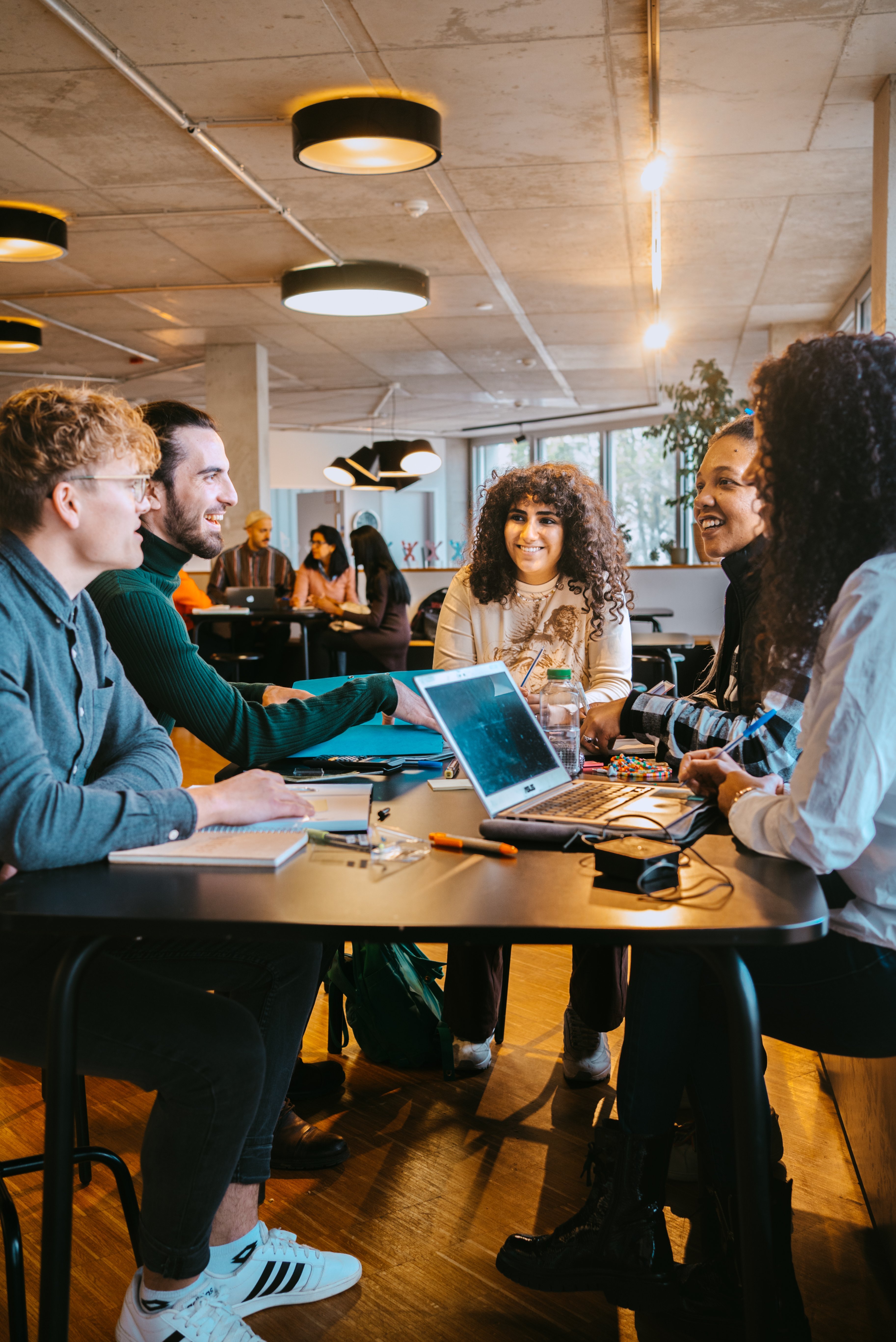 A group of five people is sitting at a table, surrounded by laptops and notebooks. They are discussing and collaborating in a modern, bright room with large windows and a relaxed atmosphere.