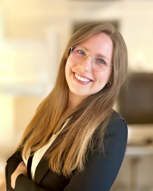 Eine lächelnde Frau mit langen, blonden Haaren und Brille trägt einen schwarzen Blazer und steht in einem modernen Büro.