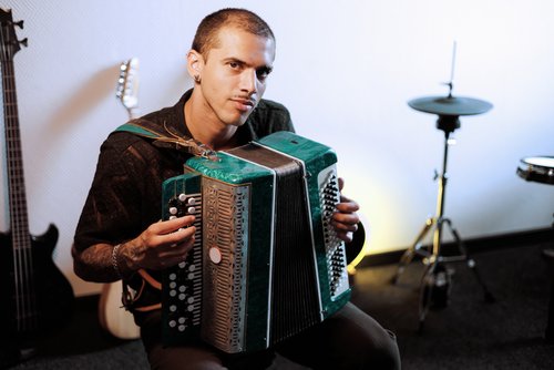 Ein Mann spielt ein Akkordeon, sitzt in einem Musikraum mit Instrumenten im Hintergrund. Er trägt ein schwarzes Hemd und schaut direkt in die Kamera.